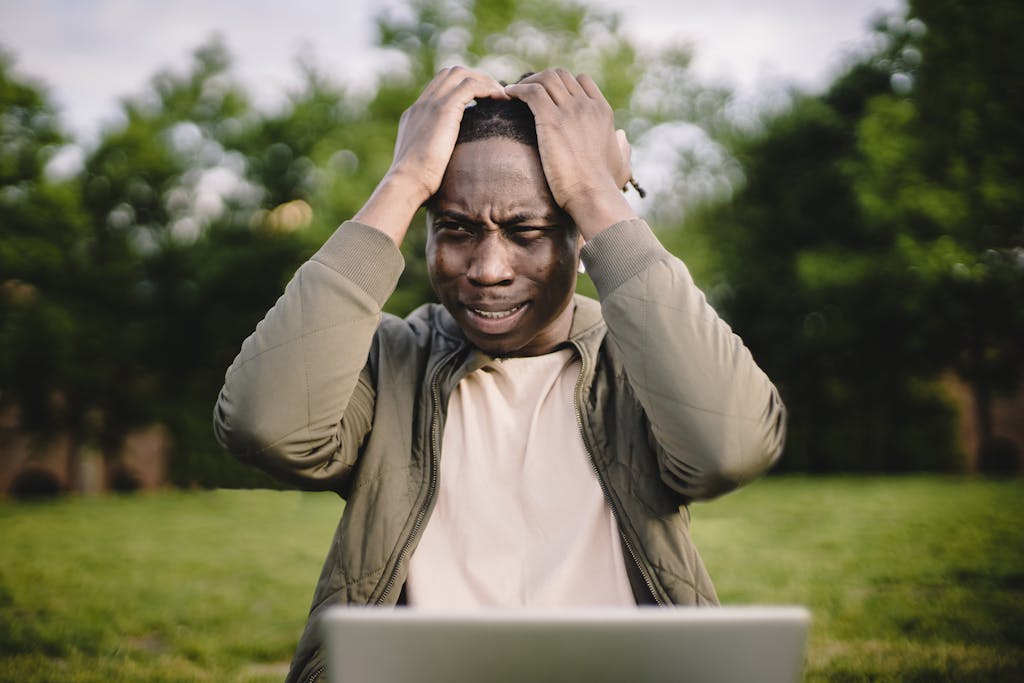 Disappointed black man holding head thinking about problem in front of laptop in park